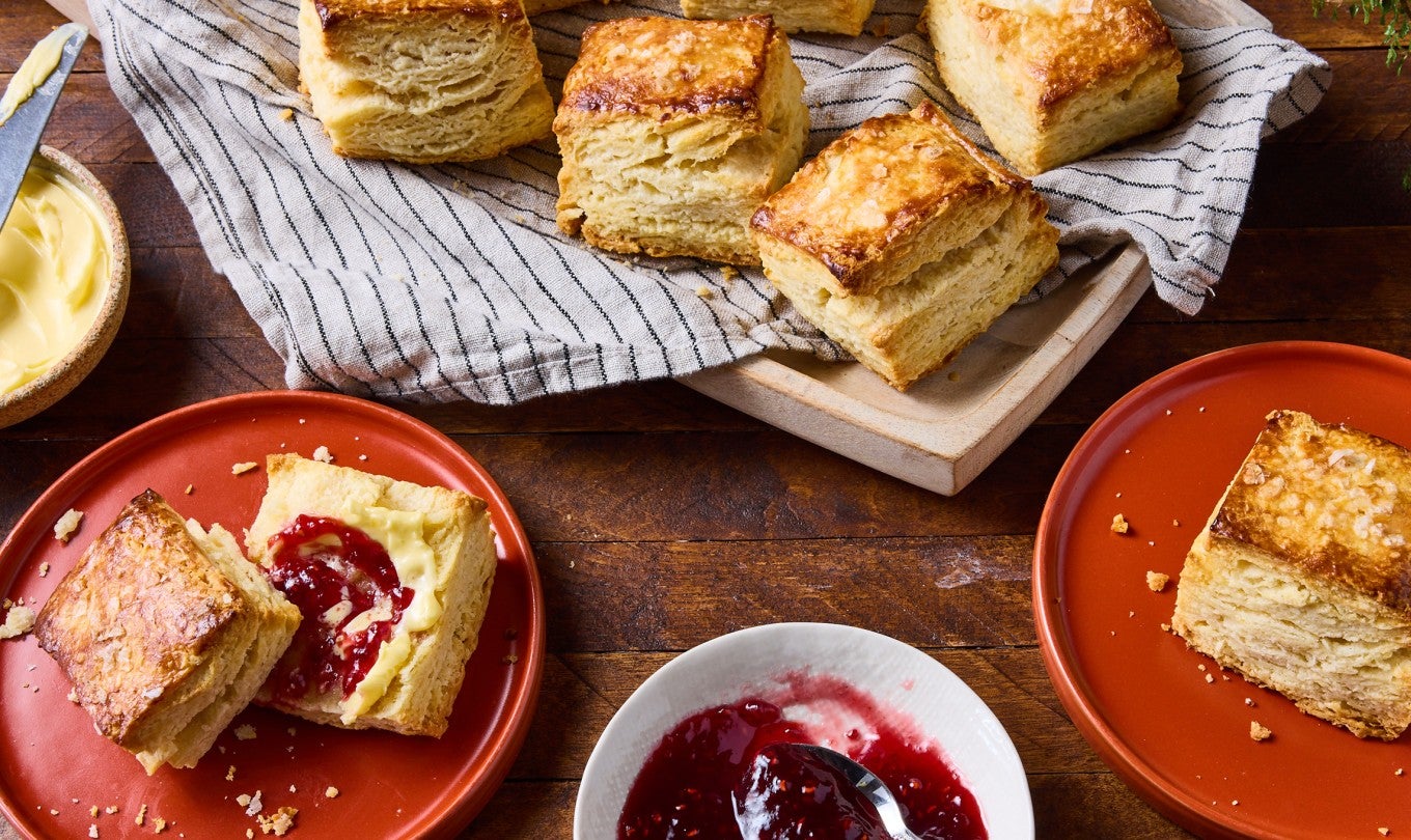 Overhead view of a sliced biscuit on a red plate and a wooden table. There is red jelly and butter on the biscuit slice.